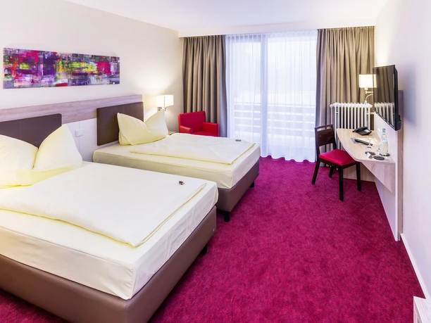 Montana Hotel Gummersbach Nord Hotelzimmer mit zwei Einzelbetten, rotem Teppich, TV, Schreibtisch und heller Fensterfront.