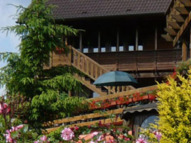 Privatzimmer Päffgen Holzhaus mit steiler Holztreppe, umgeben von blühenden Sträuchern und zahlreichen Pflanzen.