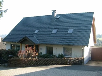 Zeitwohnen Becker Ein Einfamilienhaus mit schrägem, dunklem Dach und drei Dachfenstern, umgeben von Garten und Bäumen.