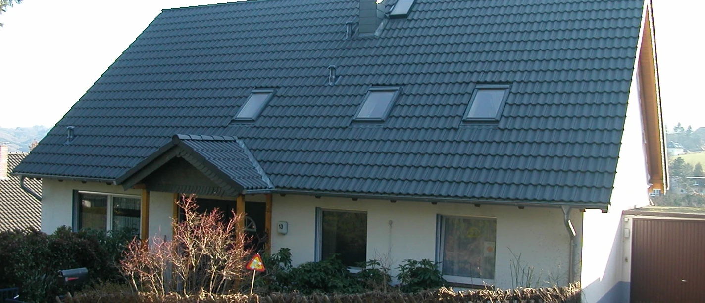 Zeitwohnen Becker Ein Einfamilienhaus mit schrägem, dunklem Dach und drei Dachfenstern, umgeben von Garten und Bäumen.