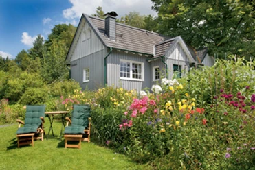 Außenansicht Gemütliches graues Holzhaus, umgeben von blühendem Garten mit Blumen, Liegestühlen und Tisch.