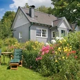 Außenansicht Gemütliches graues Holzhaus, umgeben von blühendem Garten mit Blumen, Liegestühlen und Tisch.
