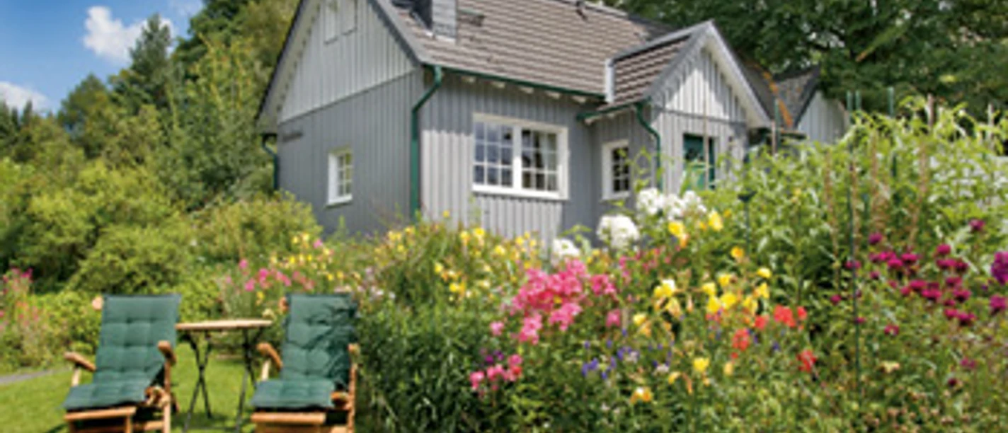 Außenansicht Gemütliches graues Holzhaus, umgeben von blühendem Garten mit Blumen, Liegestühlen und Tisch.