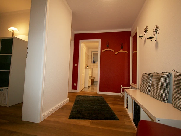 Badezimmer Flur eines Hauses mit Holzboden, einer roten Wand, Sitzbank, Spiegel und Haken. Schlafzimmer im Hintergrund.