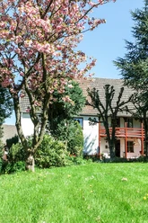 Hoffmanns Hof Zweistöckiges weißes Haus mit rotem Balkon, umgeben von blühenden Kirschbäumen und grünem Rasen.