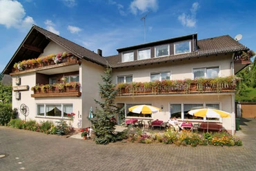 Außenansicht Die Fassade eines zweistöckigen Gästehauses mit Balkonen voller bunter Blumen und Sonnenschirmen.