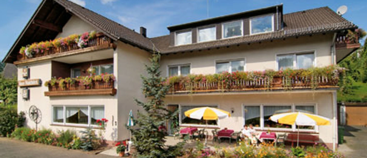 Außenansicht Die Fassade eines zweistöckigen Gästehauses mit Balkonen voller bunter Blumen und Sonnenschirmen.