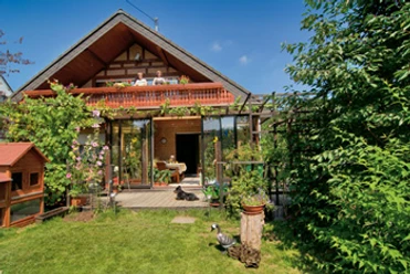 Außenansicht Ein idyllisches Landhaus mit Glasveranda, üppigem Garten und blühenden Pflanzen an einem sonnigen Tag.
