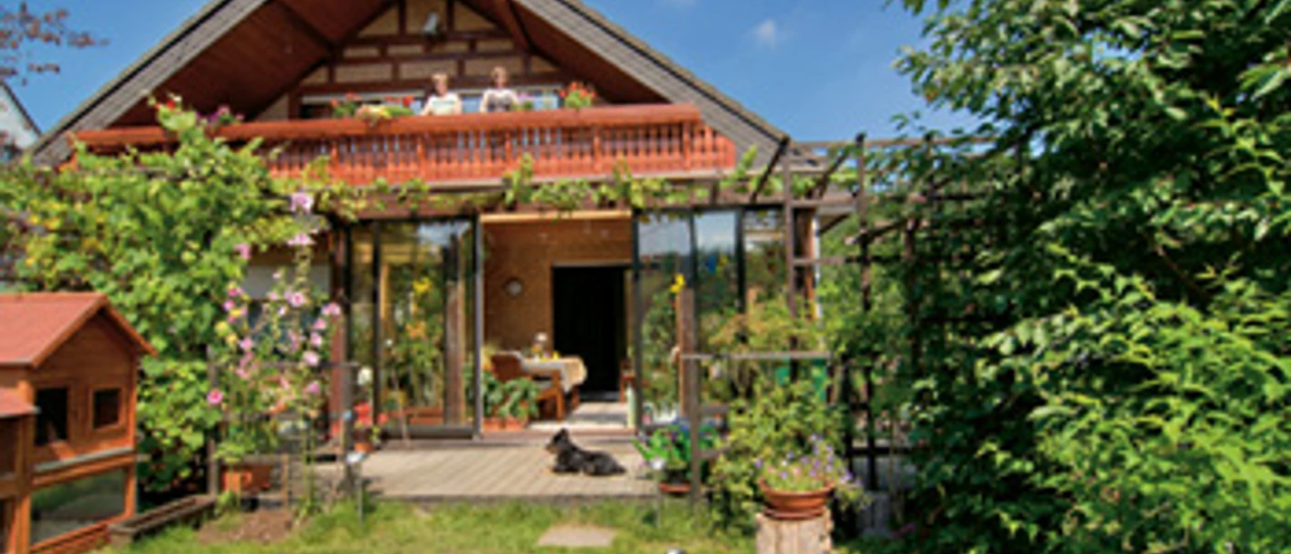 Außenansicht Ein idyllisches Landhaus mit Glasveranda, üppigem Garten und blühenden Pflanzen an einem sonnigen Tag.