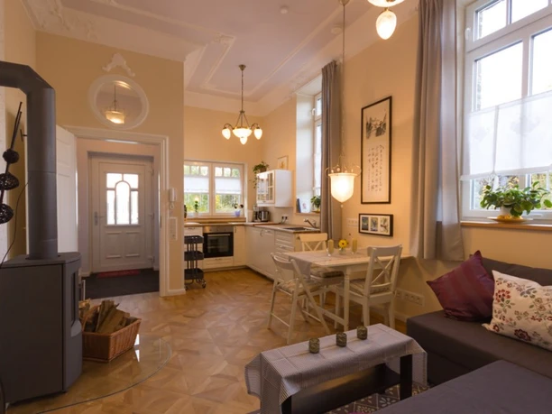 Villa Ruth - Wohnung 6 Wohnzimmer Gemütlicher Wohnraum mit Holzofen, Essbereich und moderner Küche, helle Fenster mit Vorhängen.