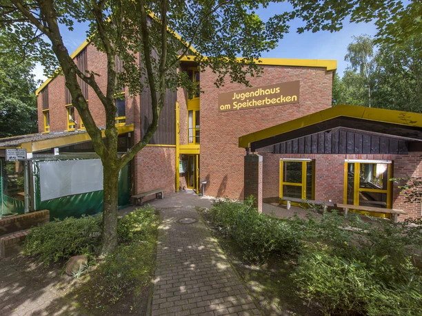 au-enansicht-2 Backsteinbau mit gelben Fensterrahmen und Eingangsbereich, umgeben von Bäumen und Grünflächen