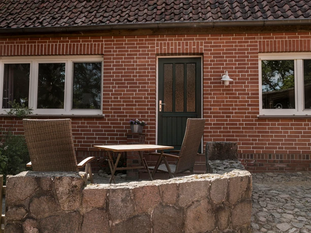 Außenansicht Rote Backsteinfassade mit gepflasterter Terrasse, Gartentisch, zwei Stühlen und einer Steinmauer.