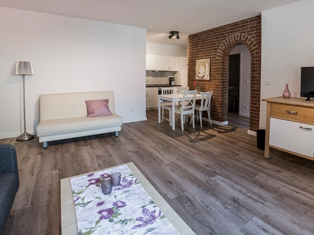 Essecke mit offener Küche Heller Wohnbereich mit Sofa, weißem Esstisch, offener Küche und einem markanten Backsteinbogen.