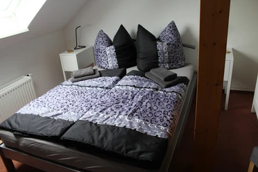 Een uitnodigende slaapkamer met een groot bed in donkere en lichte paarstinten, versierd met donzige kussens, grenzend aan een bovenverdieping met een schuin dak.