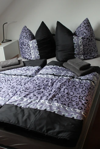 Ein einladendes Schlafzimmer mit einem großen Bett in dunklen und hellen Lilatönen, verziert mit flauschigen Kissen, angrenzend an eine obere Etage mit Dachschräge.