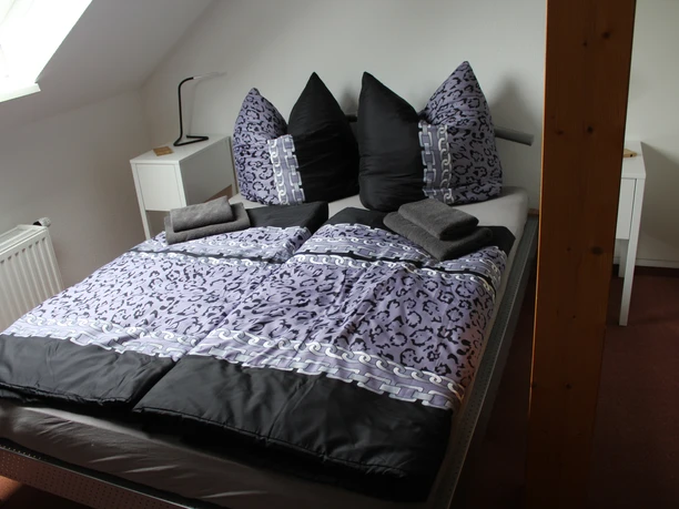 An inviting bedroom with a large bed in dark and light shades of purple, adorned with fluffy pillows, adjoining an upper floor with a sloping roof.