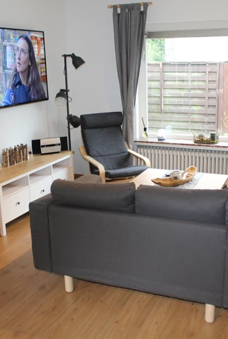 Ein moderner Wohnbereich mit grauem Sofa, Sessel, Flachbildschirm und großen Fenstern.