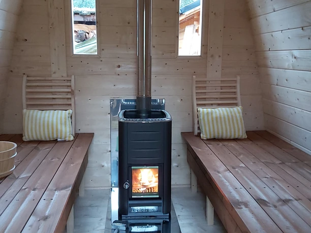 Cozy wooden hut with wood stove, two benches and striped cushions; window in the background.