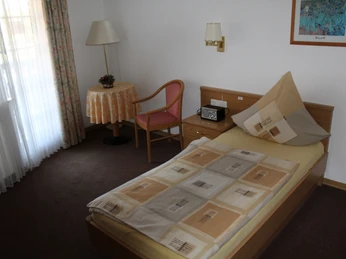 Ferienhof Meyer Einzelzimmer mit Einzelbett, Tisch, Stuhl, Nachttisch, Lampe und Fenster mit Gardinen.