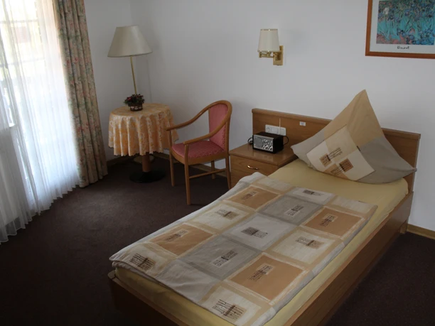 Ferienhof Meyer Einzelzimmer mit Einzelbett, Tisch, Stuhl, Nachttisch, Lampe und Fenster mit Gardinen.