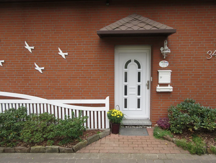 Eine weiße Eingangstür in einer Backsteinfassade, umgeben von Pflanzen und einem dekorativen Zaun.