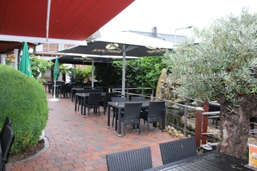 Spacious terrace with black chairs and tables under white parasols and a green backdrop.