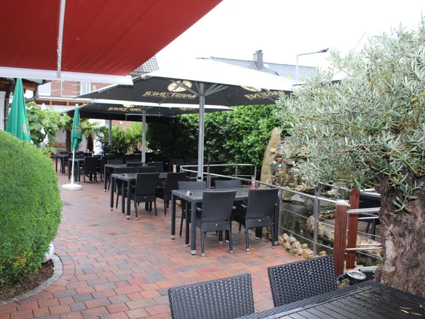 Spacious terrace with black chairs and tables under white parasols and a green backdrop.