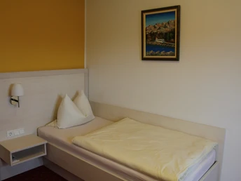 Einzelbett in einem hellen Zimmer mit gelber Wand und Bild, ausgestattet mit Nachttisch und Lampe.Single bed in a bright room with yellow wall and picture, equipped with bedside table and lamp.Enkeltseng i et lyst værelse med gul væg og billede, udstyret med natbord og lampe.Eenpersoonsbed in een lichte kamer met gele muur en foto, uitgerust met nachtkastje en lamp.
