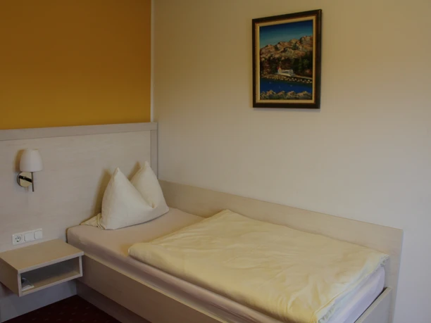 Single bed in a bright room with yellow wall and picture, equipped with bedside table and lamp.