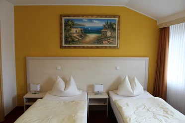 Hotel-Restaurant Platon Zwei Einzelbetten mit weißen Bettdecken in einem Zimmer mit gelb gestrichener Wand und Küstenbild.
