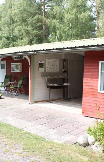 Ein rotes Holzgebäude in einer bewaldeten Umgebung mit überdachtem Sitzbereich und Grünflächen.