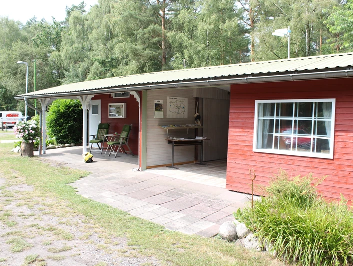 Ein rotes Holzgebäude in einer bewaldeten Umgebung mit überdachtem Sitzbereich und Grünflächen.
