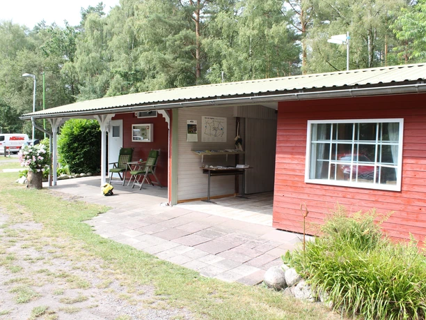 Moorcamp Ein rotes Holzgebäude in einer bewaldeten Umgebung mit überdachtem Sitzbereich und Grünflächen.