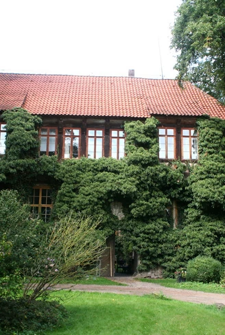 Historisches Herrenhaus mit roter Ziegelbedachung und von Efeu überwucherten Fassaden. Umgeben von üppigem Grün.
