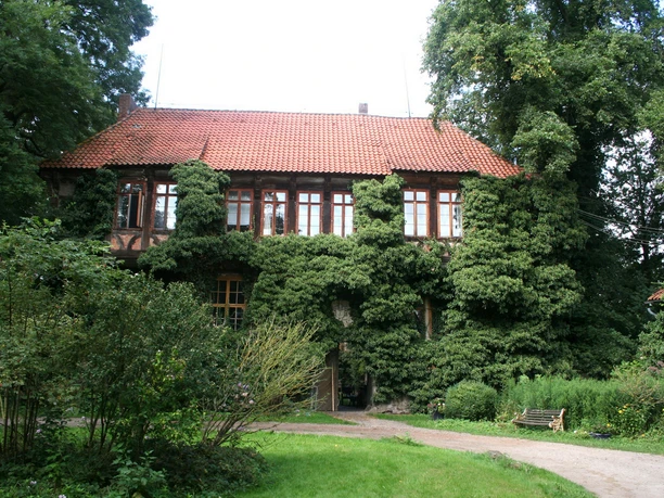 Historic manor house with red tiled roof and facades overgrown with ivy. Surrounded by lush greenery.