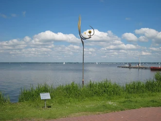 "Tanz der Winde" von Jörg Wiele Metal sculpture by Jörg Wiele on the Steinhuder Meer, with sailing boats and a cloudy sky in the background.