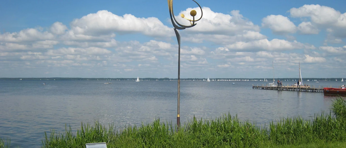 "Tanz der Winde" von Jörg Wiele Metal sculpture by Jörg Wiele on the Steinhuder Meer, with sailing boats and a cloudy sky in the background.