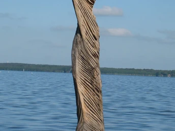 Holzskulptur "Papagena im Wind" von Klaus Wolf Simon, ragt kraftvoll aus dem ruhigen Wasser hervor.Wooden sculpture "Papagena in the wind" by Klaus Wolf Simon, juts out powerfully from the calm water.Træskulpturen "Papagena in the wind" af Klaus Wolf Simon stikker kraftfuldt ud fra det rolige vand.Houten sculptuur "Papagena in de wind" van Klaus Wolf Simon, steekt krachtig uit het kalme water.
