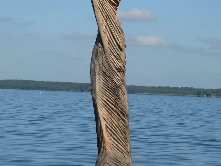 "Papagena im Wind" von Klaus Wolf Simon Holzskulptur "Papagena im Wind" von Klaus Wolf Simon, ragt kraftvoll aus dem ruhigen Wasser hervor.Wooden sculpture "Papagena in the wind" by Klaus Wolf Simon, juts out powerfully from the calm water.Træskulpturen "Papagena in the wind" af Klaus Wolf Simon stikker kraftfuldt ud fra det rolige vand.Houten sculptuur "Papagena in de wind" van Klaus Wolf Simon, steekt krachtig uit het kalme water.