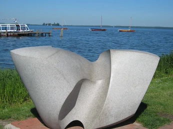"Swingende Welle" von Werner C. Sauer Eine moderne Steinskulptur mit geschwungenen Linien steht vor einem See, grasbedeckte Umgebung.A modern stone sculpture with curved lines stands in front of a lake, surrounded by grass.En moderne stenskulptur med buede linjer står foran en sø omgivet af græs.Een moderne stenen sculptuur met gebogen lijnen staat voor een meer, omringd door gras.