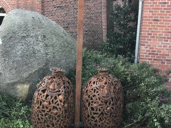 Lieblingsort Hengstforder Mühle Auf dem Bild sind zwei kunstvoll durchbrochene Metallstrukturen in Form großer Mehlsäcke zu sehen, die neben einem hohen Metallpfahl mit einer blauen Kugel an der Spitze stehen. Im Hintergrund ist eine hohe Mauer aus roten Ziegelsteinen zu erkennen.
