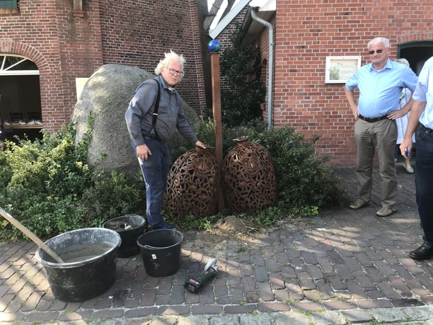 Künstler Jörg Ridderbusch beim Aufbau Künstler Jörg Ridderbusch steht neben zwei rostfarbenen Skulpturen im Freien, umgeben von Ziegelsteinwänden und Pflanzen. Werkzeuge und Schalen liegen in der Nähe auf dem Kopfsteinpflaster.