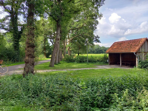 Schutzhütte Marschstrasse Schutzhütte an einem mit Bäumen gesäumten Weg, umgeben von grüner Landschaft und blauem Himmel.