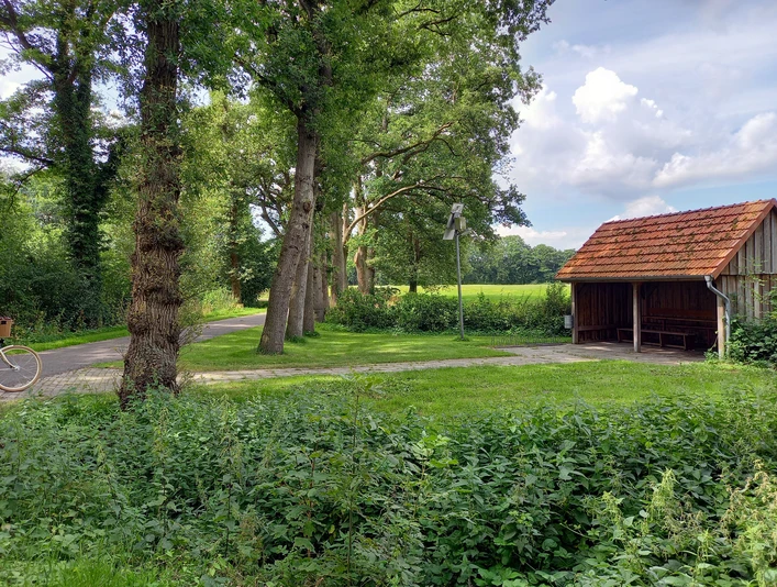 Schutzhütte Marschstrasse Schutzhütte an einem mit Bäumen gesäumten Weg, umgeben von grüner Landschaft und blauem Himmel.