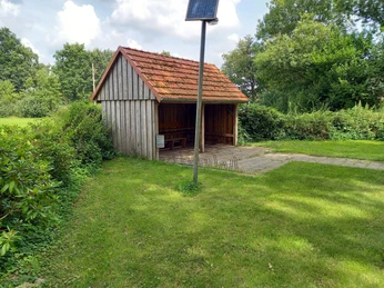 Rastplatz Apermarsch Ein Rastplatz inmitten grüner Vegetation mit einem überdachten Holzunterstand auf einer Rasenfläche. Ein Solarmast spendet Licht.