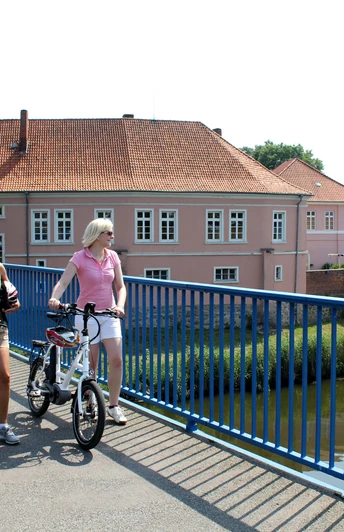 Zwei Frauen auf Fahrrädern überqueren eine Brücke vor dem historischen Grafenschloss in Hoya.