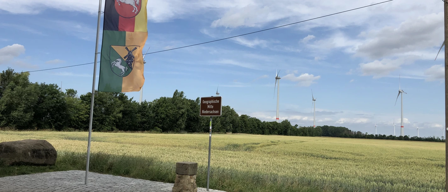 Open space with flags, drive to the wind turbine, surrounded by fields and wooded horizon.
