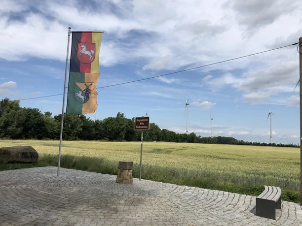 Open space with flags, drive to the wind turbine, surrounded by fields and wooded horizon.