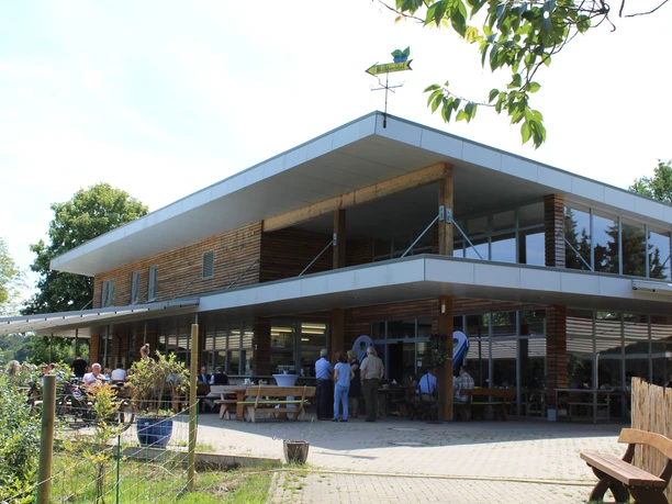 Large wooden building with people on the terrace. Garden chairs, tables and trees in the foreground.