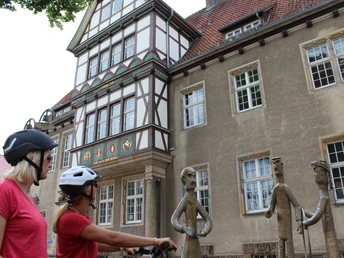 Zwei Radfahrerinnen mit Helmen betrachten moderne Bronzefiguren vor einem Fachwerkgebäude.Two cyclists with helmets look at modern bronze figures in front of a half-timbered building.To cyklister med hjelm kigger på moderne bronzefigurer foran en bindingsværksbygning.Twee fietsers met helmen kijken naar moderne bronzen figuren voor een vakwerkgebouw.